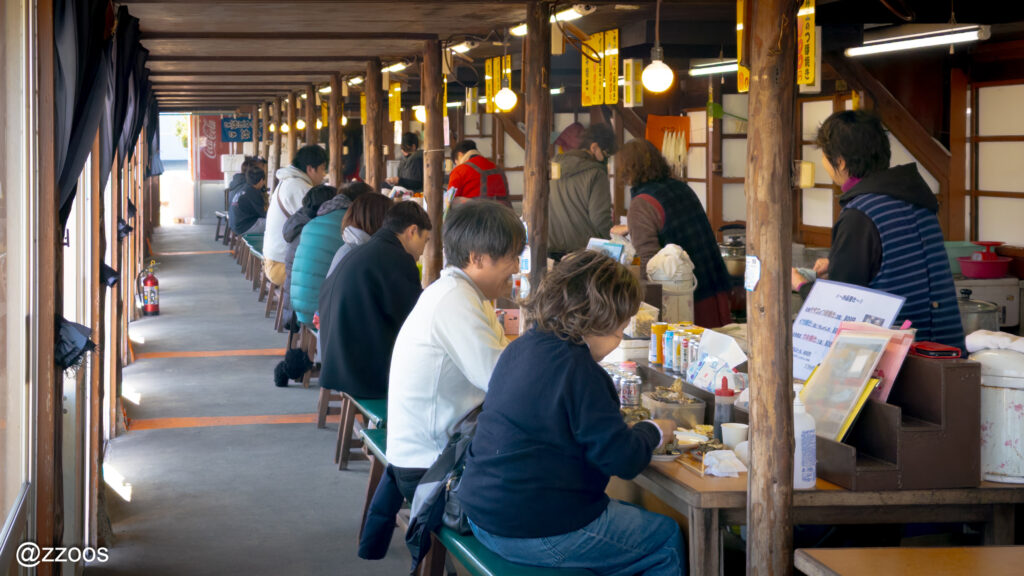 People are sitting at a wooden stall inside a timber building eating grilled turban shells.